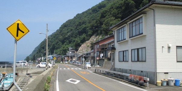 海沿いの道と住宅が並ぶ風景、右分岐の道路標識と山が背景に広がる日本の沿岸地域のイメージ